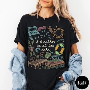 Puede incluir: Camiseta negra con un dise&ntilde;o gr&aacute;fico colorido con ilustraciones tem&aacute;ticas de lago, que incluyen una nevera port&aacute;til, gafas de sol, protector solar, un sol y un salvavidas. Tambi&eacute;n se incluye el texto "I'd rather be at the lake".