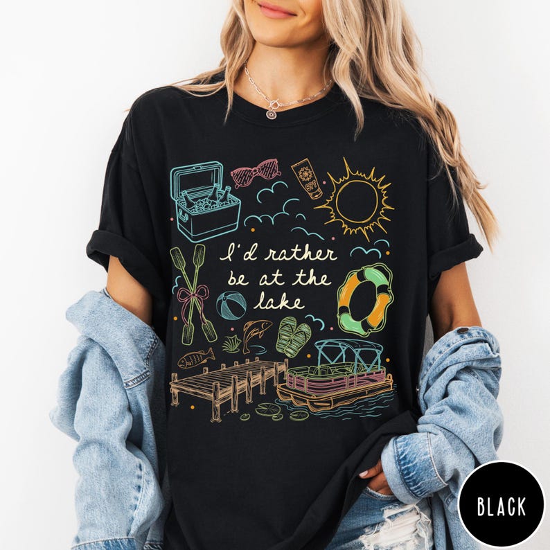Puede incluir: Camiseta negra con un dise&ntilde;o gr&aacute;fico colorido con ilustraciones tem&aacute;ticas de lago, que incluyen una nevera port&aacute;til, gafas de sol, protector solar, un sol y un salvavidas. Tambi&eacute;n se incluye el texto "I'd rather be at the lake".