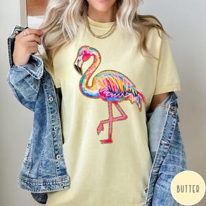 May include: A light yellow t-shirt featuring a colorful flamingo design. The flamingo has a rainbow-colored body, pink legs, and a black beak. The shirt is a casual style, suitable for everyday wear.