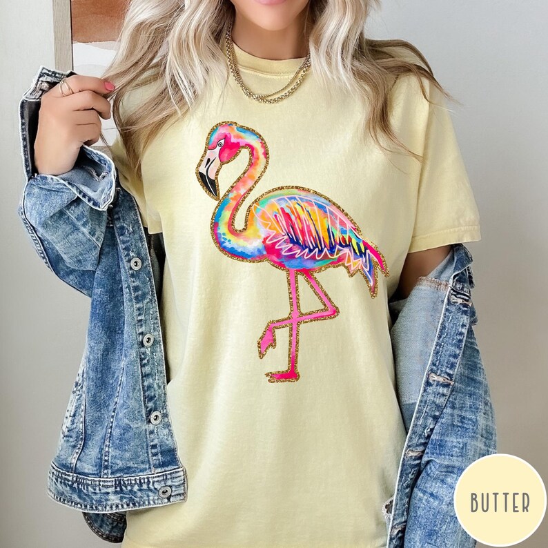 May include: A light yellow t-shirt featuring a colorful flamingo design. The flamingo has a rainbow-colored body, pink legs, and a black beak. The shirt is a casual style, suitable for everyday wear.