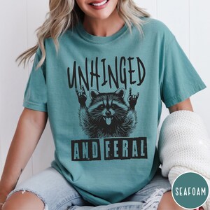 May include: A teal blue t-shirt with a black graphic of a raccoon with its arms raised in a rock and roll hand gesture. The text "Unhinged and Feral" is printed below the raccoon.
