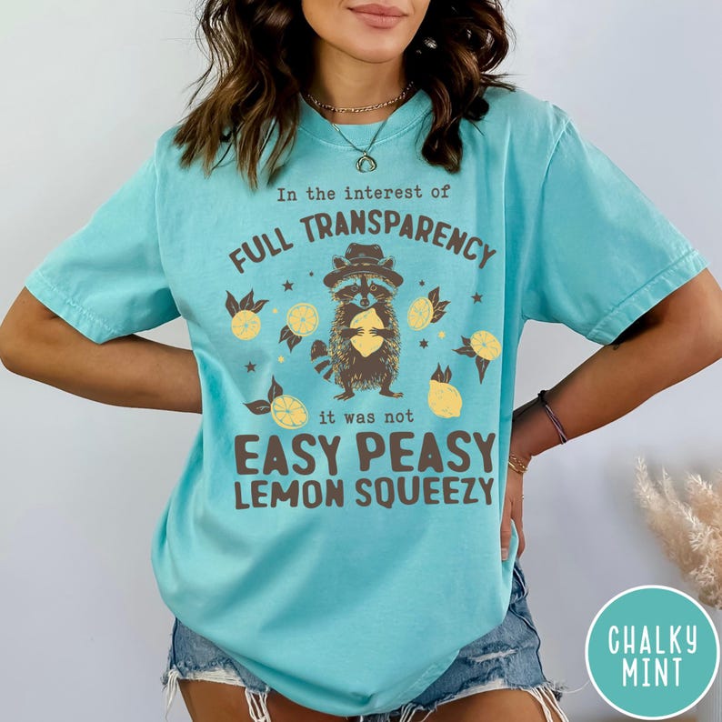 May include: A turquoise blue t-shirt with a graphic of a raccoon wearing a hat and holding a lemon. The text on the shirt reads "In the interest of FULL TRANSPARENCY it was not EASY PEASY LEMON SQUEEZY".