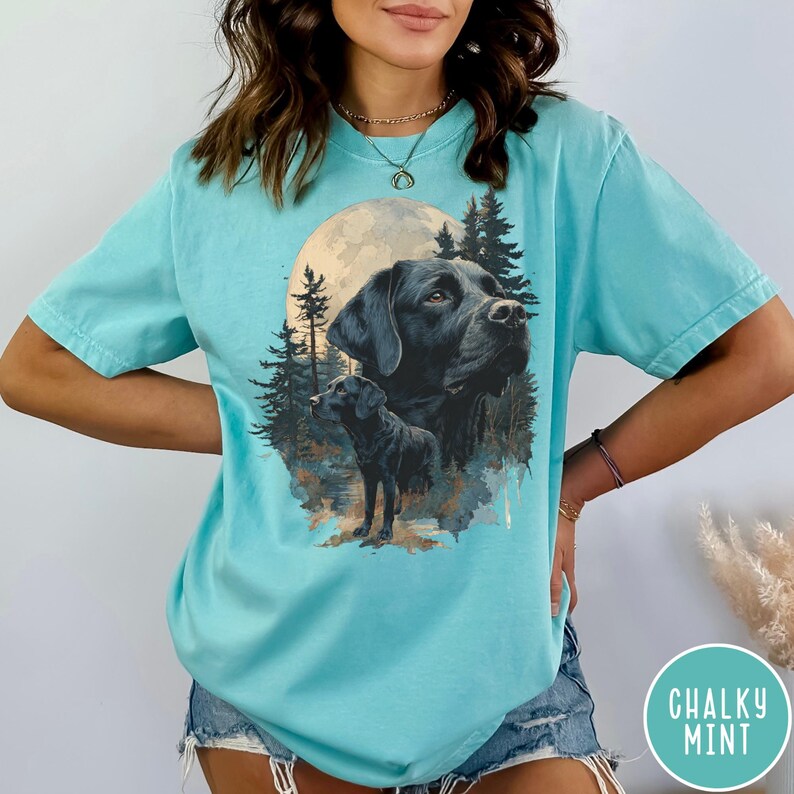 May include: A light blue t-shirt featuring a graphic of a black Labrador Retriever with a full moon and forest background. The shirt has a relaxed fit and short sleeves. The text "CHALKY MINT" is in a circle on the bottom right.