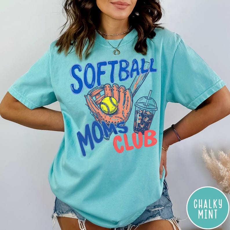 May include: A light blue t-shirt with the words "SOFTBALL MOMS CLUB" in blue and coral lettering. The shirt features a softball glove, a yellow softball, and a drink with ice. The shirt is a comfortable, casual style.