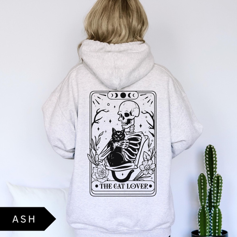 May include: A gray hoodie with a black and white illustration of a skeleton holding a black cat. The illustration is surrounded by a black frame with the text "The Cat Lover" at the bottom.