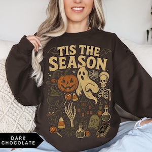 Puede incluir: Sudadera de color chocolate oscuro con un dise&ntilde;o gr&aacute;fico de tem&aacute;tica de Halloween. El dise&ntilde;o incluye una calabaza, un fantasma, un esqueleto y la frase "Tis the Season" en letras doradas. Tambi&eacute;n se incluyen elementos adicionales de Halloween como un sombrero de bruja, un caldero y caramelos.