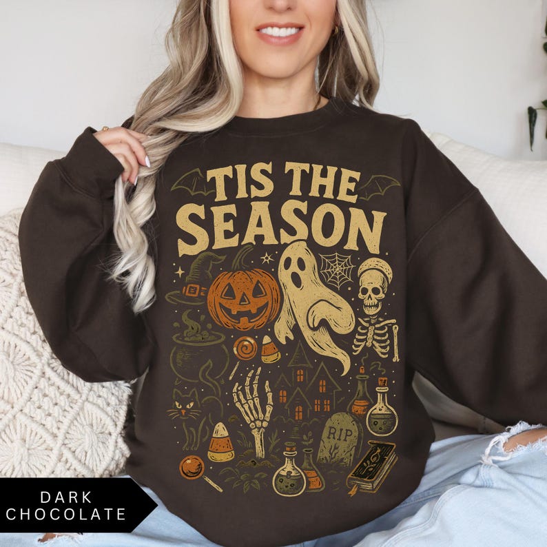 Puede incluir: Sudadera de color chocolate oscuro con un dise&ntilde;o gr&aacute;fico de tem&aacute;tica de Halloween. El dise&ntilde;o incluye una calabaza, un fantasma, un esqueleto y la frase "Tis the Season" en letras doradas. Tambi&eacute;n se incluyen elementos adicionales de Halloween como un sombrero de bruja, un caldero y caramelos.