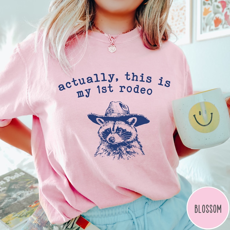 May include: Pink t-shirt with the text "actually, this is my 1st rodeo" and a cartoon raccoon wearing a cowboy hat.