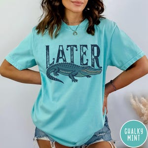 May include: A light blue t-shirt with the word "LATER" in dark blue block letters above a detailed illustration of a blue alligator. The shirt is a solid color and has short sleeves.