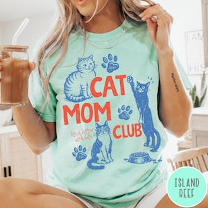 K&ouml;nnte beinhalten: Mintgr&uuml;nes T-Shirt mit den Worten "CAT MOM CLUB" in Rot und Blau. Das Shirt zeigt Illustrationen von Katzen und Pfotenabdr&uuml;cken. Der Text "life is better with cats" ist ebenfalls enthalten.