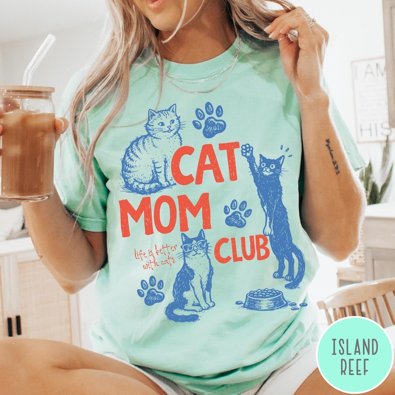K&ouml;nnte beinhalten: Mintgr&uuml;nes T-Shirt mit den Worten "CAT MOM CLUB" in Rot und Blau. Das Shirt zeigt Illustrationen von Katzen und Pfotenabdr&uuml;cken. Der Text "life is better with cats" ist ebenfalls enthalten.