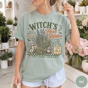 May include: A sage green t-shirt featuring a graphic print with the text "Witch's Herb Garden" and illustrations of herbs, a mushroom, a potion bottle, and a crescent moon. The design includes the phrases "Freshly Gathered," "Locally Grown Herbs," and "Moonlit Grown."