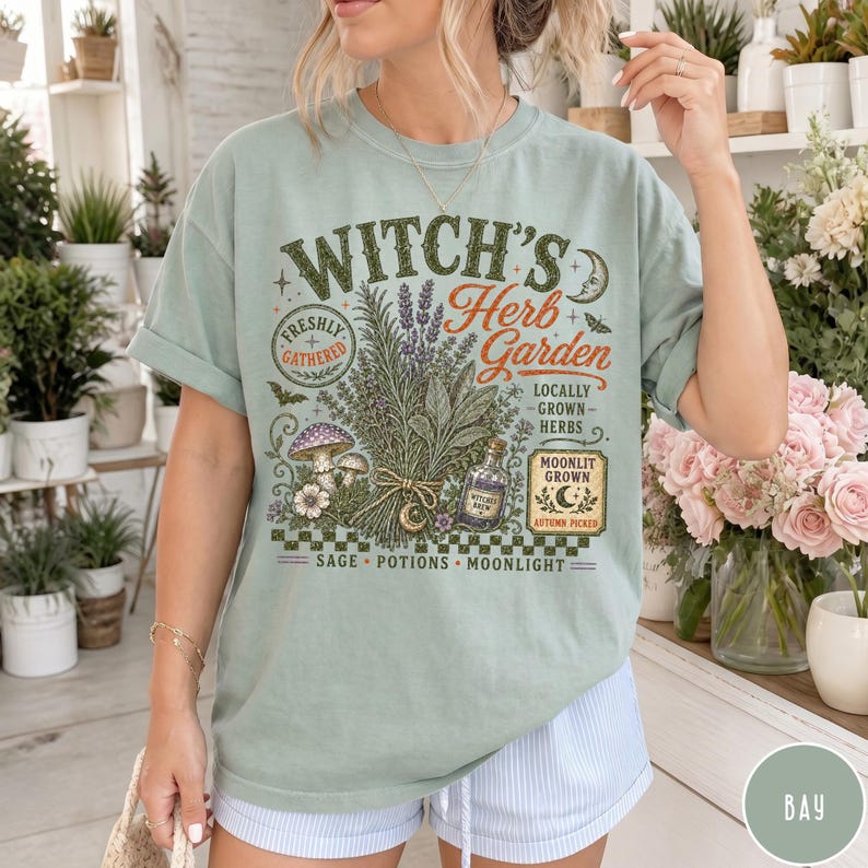 May include: A sage green t-shirt featuring a graphic print with the text "Witch's Herb Garden" and illustrations of herbs, a mushroom, a potion bottle, and a crescent moon. The design includes the phrases "Freshly Gathered," "Locally Grown Herbs," and "Moonlit Grown."