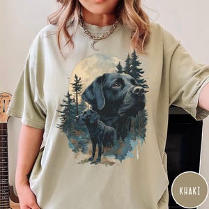 May include: Khaki-colored t-shirt featuring a graphic of a black Labrador Retriever with a full moon and forest background. The design includes two images of the dog, one a close-up of the face and the other a full body shot. The shirt is a relaxed fit.