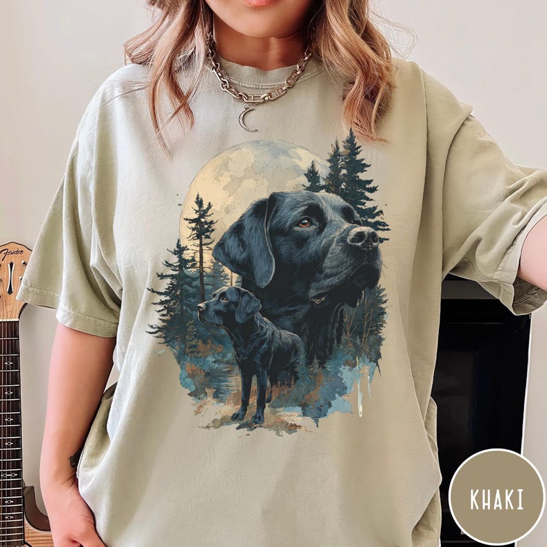 May include: Khaki-colored t-shirt featuring a graphic of a black Labrador Retriever with a full moon and forest background. The design includes two images of the dog, one a close-up of the face and the other a full body shot. The shirt is a relaxed fit.