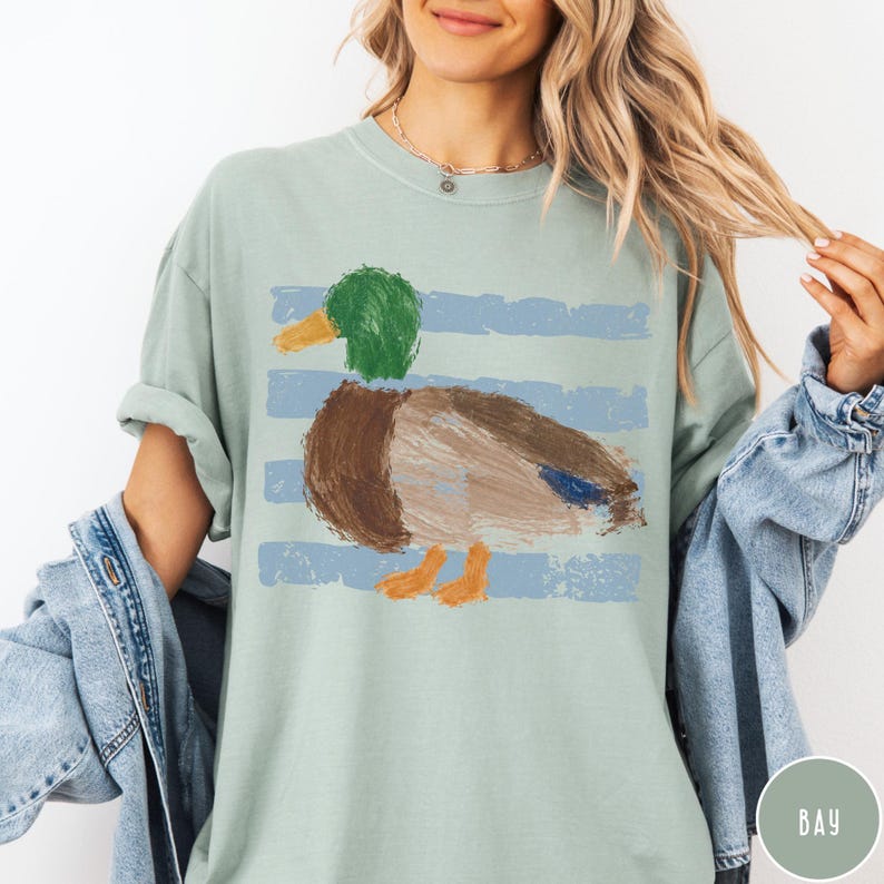 May include: A sage green short-sleeve t-shirt featuring a graphic of a mallard duck with a green head, brown body, and orange feet. The duck is layered over horizontal light blue brushstroke stripes. The shirt has a crew neckline.