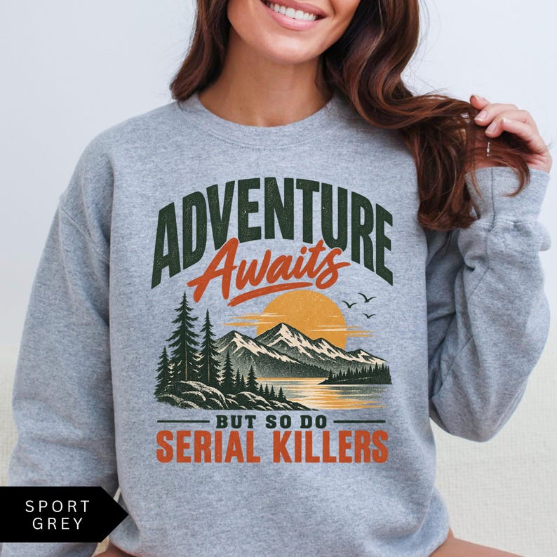 May include: A sport grey crewneck sweatshirt featuring a graphic print of a mountain landscape with a sunset, pine trees, and birds. The text reads "ADVENTURE Awaits BUT SO DO SERIAL KILLERS" in dark green and orange lettering on the front chest.