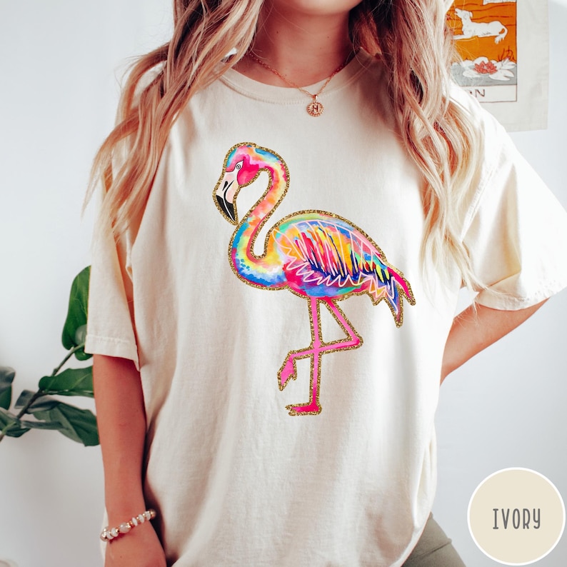 May include: Ivory-colored t-shirt featuring a vibrant, watercolor-style flamingo design. The flamingo is depicted in shades of pink, yellow, blue, and green, with gold glitter accents. The shirt has a relaxed fit and short sleeves.