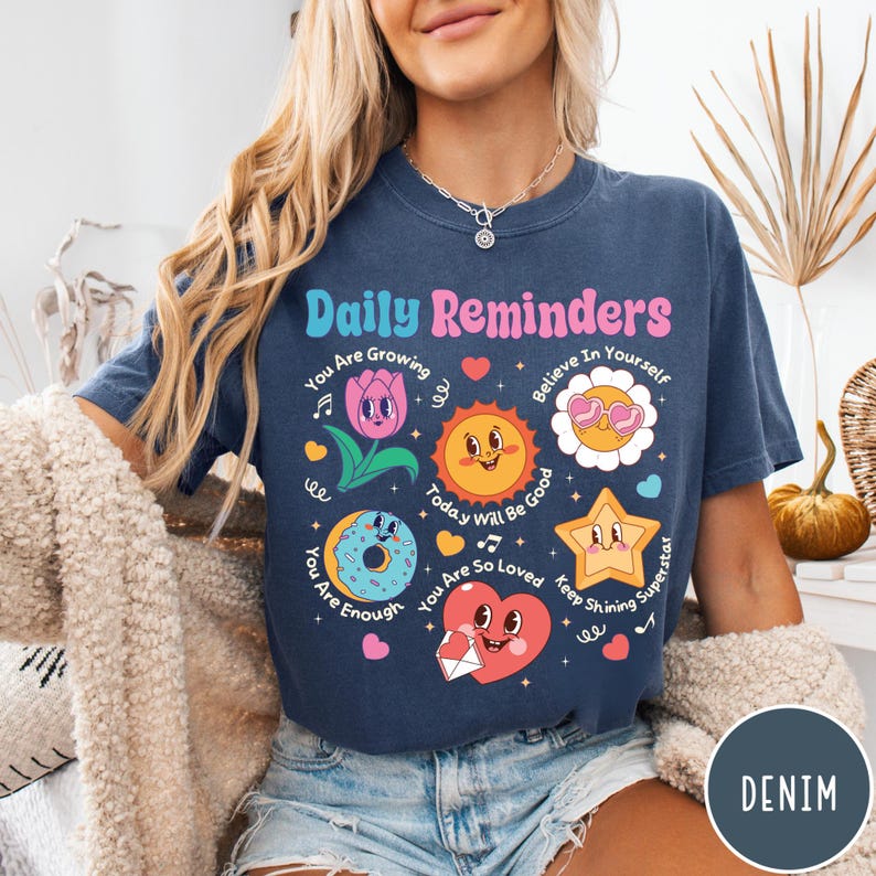 May include: A navy blue t-shirt with the words "Daily Reminders" in a retro font. The shirt features colorful illustrations of a flower, sun, donut, star, and heart with positive affirmations. The word "DENIM" is in a circle on the bottom right.