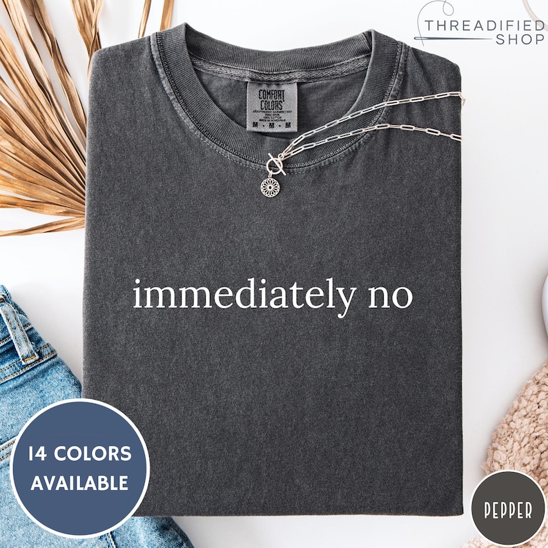 May include: Dark grey t-shirt with the words "immediately no" in white. The shirt has a crew neck and short sleeves. A silver necklace with a circular pendant is draped across the shirt. The shirt is folded and displayed on a white surface.