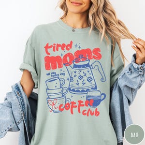 May include: A light green t-shirt with the words "tired moms coffee club" in red and blue. The design includes a teapot, stacked mugs, and the phrase "all day every day".