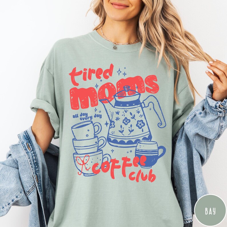 May include: A light green t-shirt with the words "tired moms coffee club" in red and blue. The design includes a teapot, stacked mugs, and the phrase "all day every day".