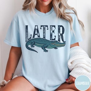 May include: Light blue t-shirt with the word "LATER" in dark blue, above a detailed illustration of a green and blue alligator. The shirt is a casual style.