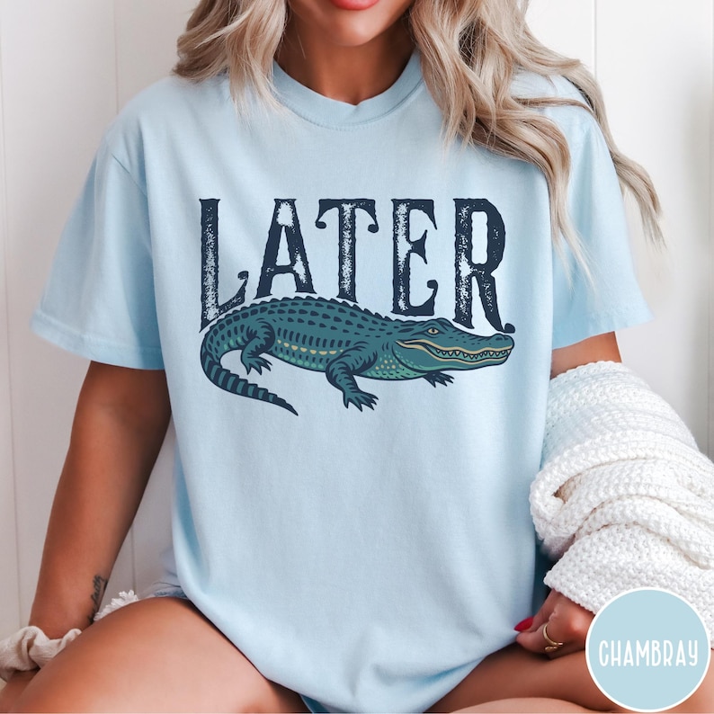 May include: Light blue t-shirt with the word "LATER" in dark blue, above a detailed illustration of a green and blue alligator. The shirt is a casual style.