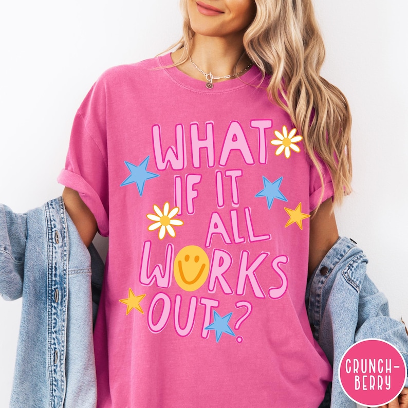 May include: A bright pink t-shirt featuring the text WHAT IF IT ALL WORKS OUT? in large pink letters. The design includes a yellow smiley face, white daisies, and blue and yellow stars scattered around the text. A blue denim jacket is layered over.