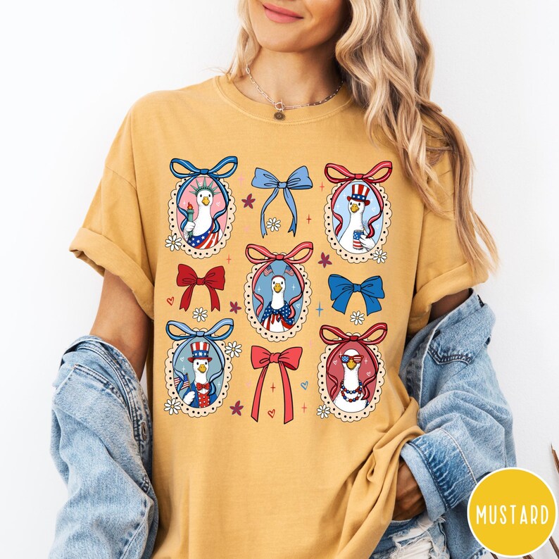May include: Mustard-colored t-shirt featuring a patriotic goose design. The design includes several goose illustrations wearing patriotic accessories, surrounded by bows and floral accents. The shirt has a relaxed fit and short sleeves.