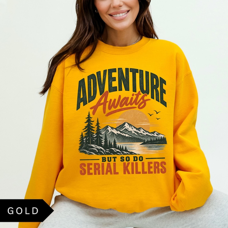 May include: A gold-colored crewneck sweatshirt featuring a graphic print of a mountain landscape with a lake, pine trees, and a sunset. The text reads ADVENTURE Awaits BUT SO DO SERIAL KILLERS in dark green, orange, and red lettering across the front.