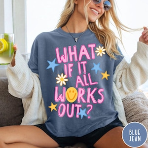 May include: A slate blue short-sleeved t-shirt featuring the pink text "WHAT IF IT ALL WORKS OUT?" with a yellow smiley face, white daisies, and blue and yellow stars printed on the front. The shirt is worn with a cream-colored cardigan.