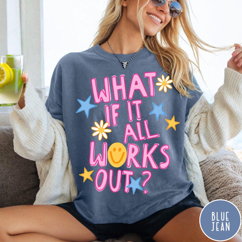 May include: A slate blue short-sleeved t-shirt featuring the pink text "WHAT IF IT ALL WORKS OUT?" with a yellow smiley face, white daisies, and blue and yellow stars printed on the front. The shirt is worn with a cream-colored cardigan.