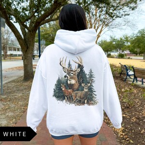 Op de afbeelding: Witte hoodie met een hertenafbeelding op de rug. De afbeelding toont een bok met grote geweien, twee kleinere herten en dennenbomen. Het woord "WHITE" staat in een zwart kader linksonder.