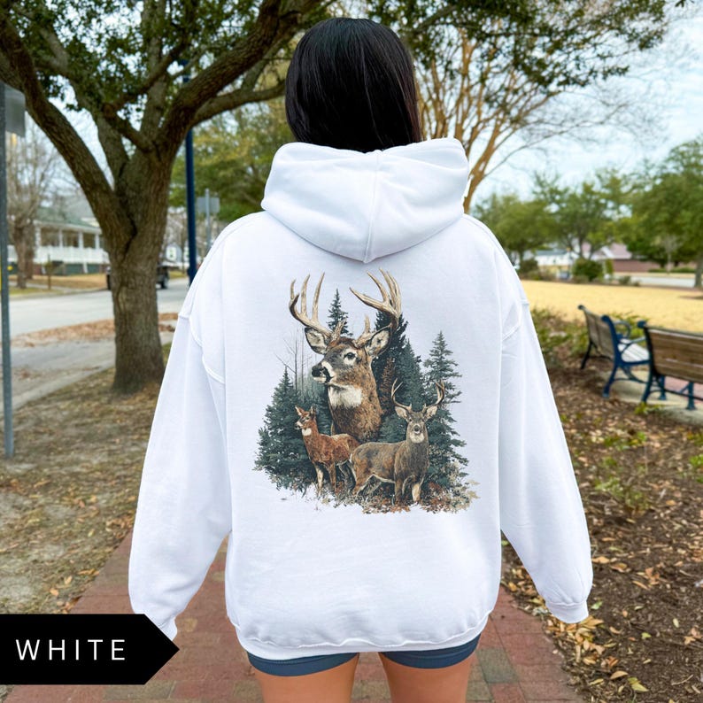 Op de afbeelding: Witte hoodie met een hertenafbeelding op de rug. De afbeelding toont een bok met grote geweien, twee kleinere herten en dennenbomen. Het woord "WHITE" staat in een zwart kader linksonder.