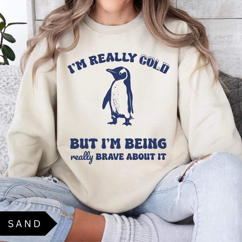 May include: A beige sweatshirt with a penguin graphic and the text "I'm really cold but I'm being really brave about it".