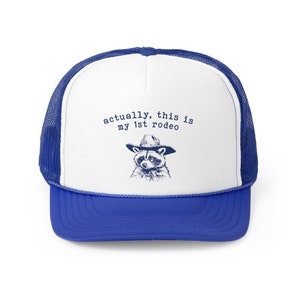 May include: A white and blue trucker hat with a blue mesh back. The front of the hat features a raccoon wearing a cowboy hat and the text "actually, this is my 1st rodeo".