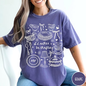 May include: A purple t-shirt featuring a white illustration of vintage tableware, such as teacups and plates, and a duck. The text "I'd rather be thrifting" is printed on the shirt.