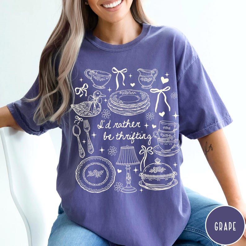 May include: A purple t-shirt featuring a white illustration of vintage tableware, such as teacups and plates, and a duck. The text "I'd rather be thrifting" is printed on the shirt.