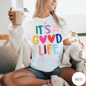 K&ouml;nnte beinhalten: Ein wei&szlig;es Kurzarm-T-Shirt mit dem bunten Schriftzug IT'S A GOOD LIFE auf der Vorderseite. Die Buchstaben sind in verschiedenen Farben wie Gr&uuml;n, Lila, Pink, Gelb, Blau und Orange gehalten, wobei zwei Herzformen den Buchstaben O ersetzen.