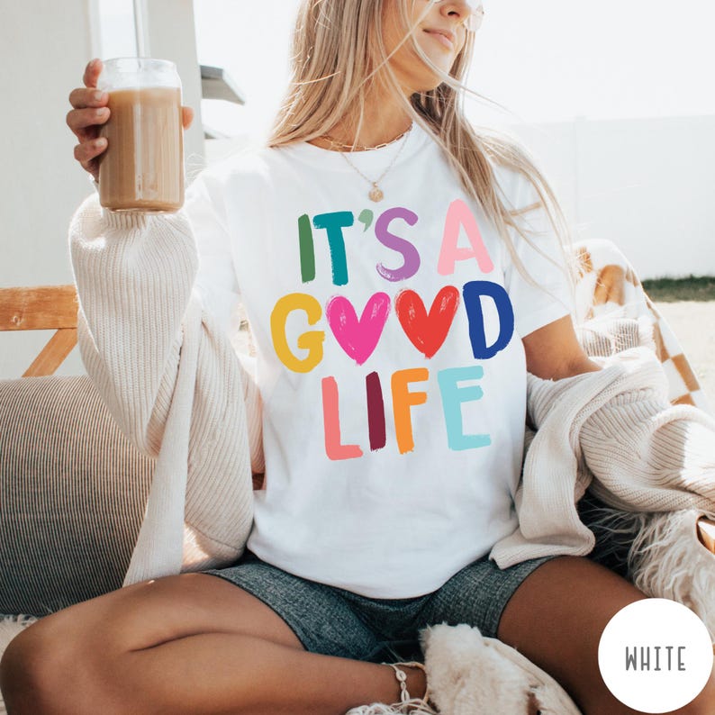 K&ouml;nnte beinhalten: Ein wei&szlig;es Kurzarm-T-Shirt mit dem bunten Schriftzug IT'S A GOOD LIFE auf der Vorderseite. Die Buchstaben sind in verschiedenen Farben wie Gr&uuml;n, Lila, Pink, Gelb, Blau und Orange gehalten, wobei zwei Herzformen den Buchstaben O ersetzen.