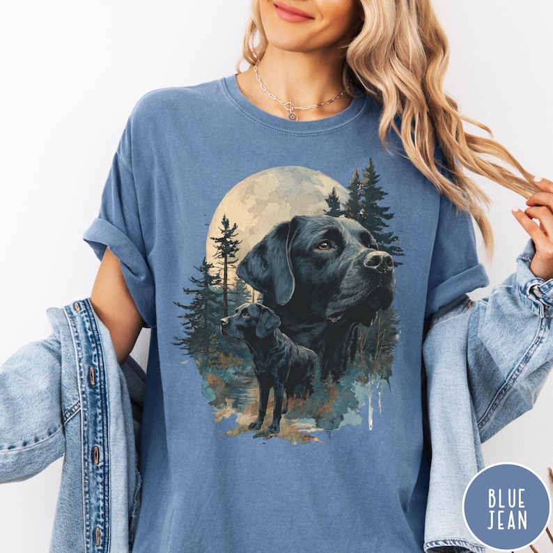 May include: Blue t-shirt featuring a graphic of two black Labrador Retrievers with a full moon and forest background. The shirt has a relaxed fit and rolled sleeves. The text "BLUE JEAN" is printed on a circular label at the bottom right.
