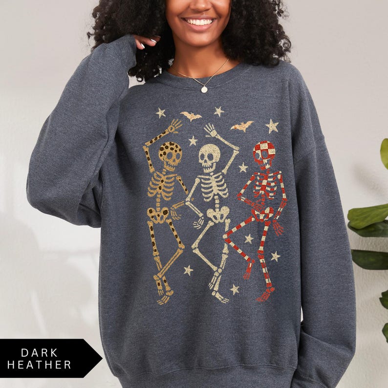 Puede incluir: Sudadera gris oscuro con un dise&ntilde;o de tres esqueletos bailando en oro, blanco y rojo. El dise&ntilde;o incluye estrellas y murci&eacute;lagos, con el texto "DARK HEATHER" en una flecha negra.