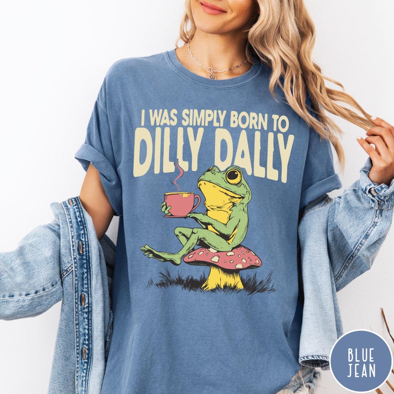 May include: Blue t-shirt with the text "I WAS SIMPLY BORN TO DILLY DALLY". The graphic shows a green frog sitting on a mushroom, holding a pink teacup. The shirt is a solid blue color.
