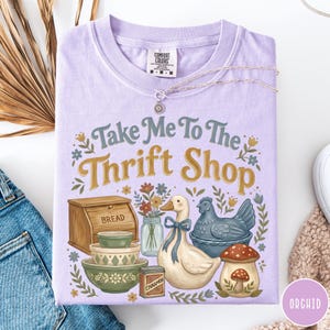 Op de afbeelding: Een lichtpaars Comfort Colors T-shirt met de tekst "Take Me To The Thrift Shop" en illustraties van een brooddoos, kommen, een eend en een paddenstoel. Een zilveren ketting ligt over het shirt.