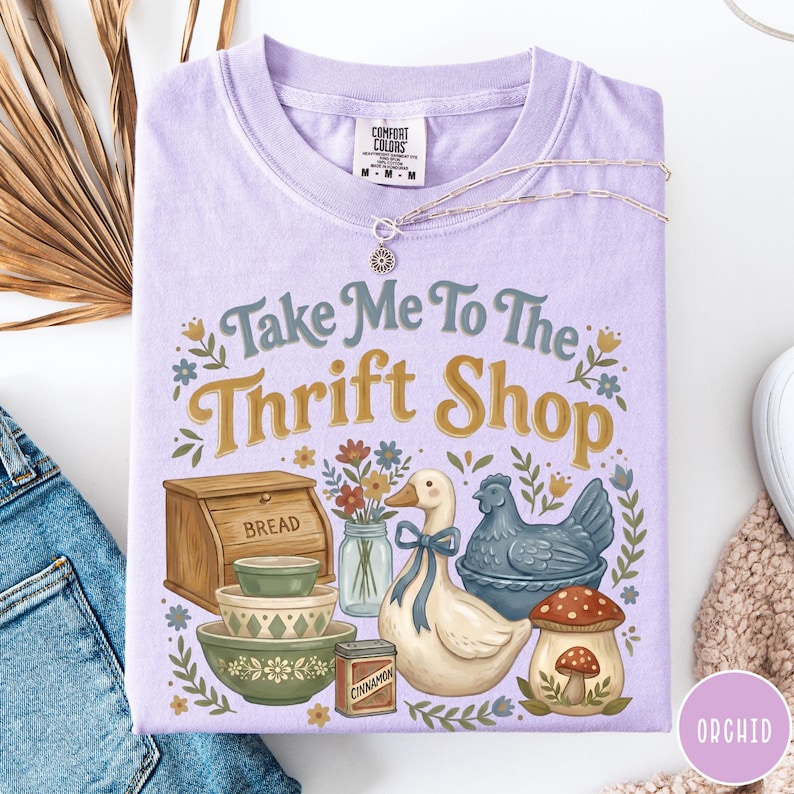 Op de afbeelding: Een lichtpaars Comfort Colors T-shirt met de tekst "Take Me To The Thrift Shop" en illustraties van een brooddoos, kommen, een eend en een paddenstoel. Een zilveren ketting ligt over het shirt.