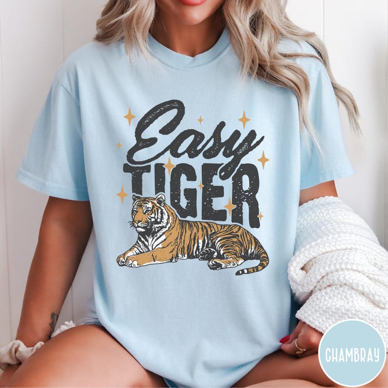 May include: A light blue t-shirt featuring a graphic of a tiger and the text "Easy Tiger" in black. The tiger is brown and black with a white background.