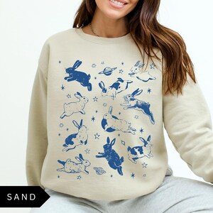 May include: A sand-coloured sweatshirt adorned with a navy blue rabbit and space-themed pattern. The design includes rabbits in various poses, with some having white markings. The word "SAND" is printed in black at the bottom.