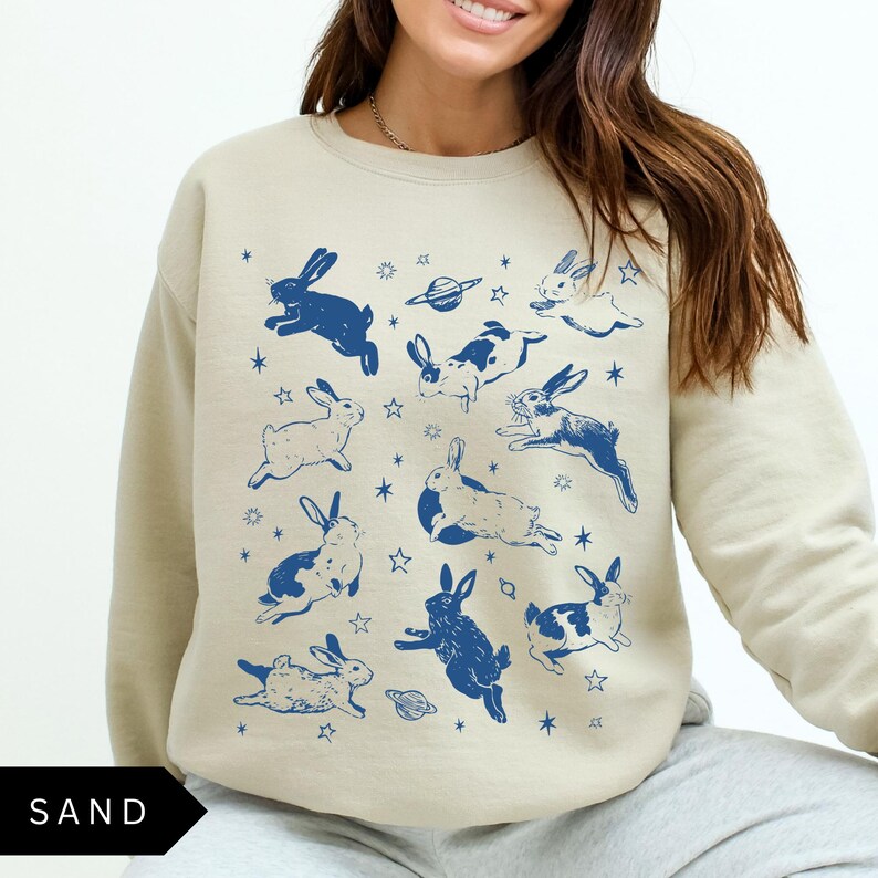 May include: A sand-coloured sweatshirt adorned with a navy blue rabbit and space-themed pattern. The design includes rabbits in various poses, with some having white markings. The word "SAND" is printed in black at the bottom.