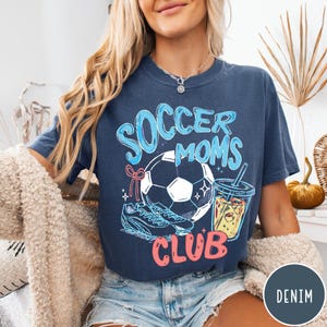 May include: A navy blue t-shirt with the words "Soccer Moms Club" in a playful font. The graphic includes a soccer ball, cleats, and a drink. The shirt is a comfortable, casual style.